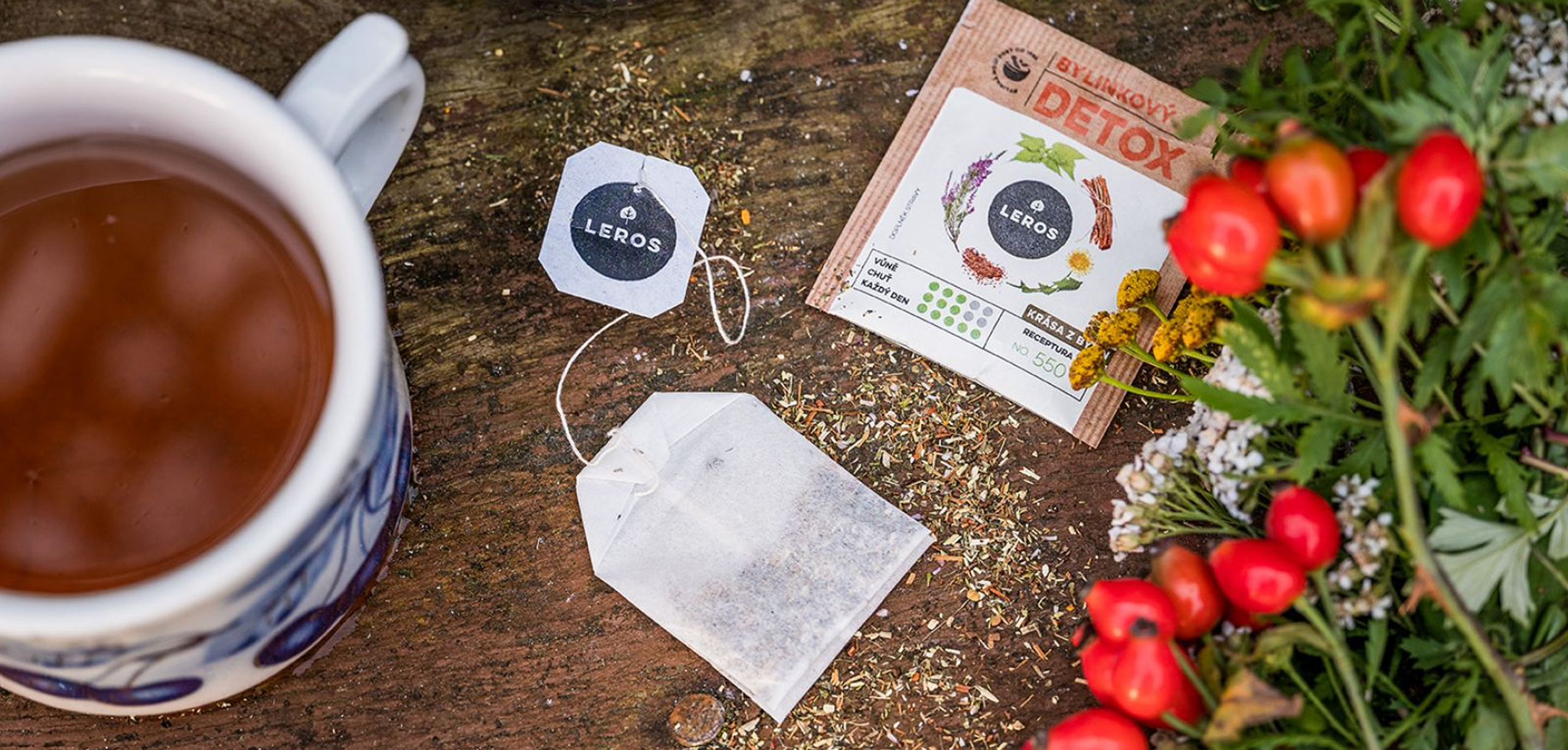 Cup of herbal tea with LEROS teabag, detox sachet and fresh red berries and herbs arranged on a rustic wooden surface