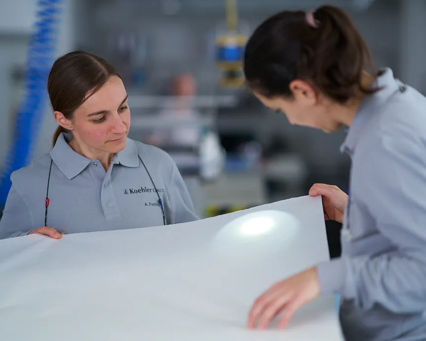 Two female technicians at Koehler plant inspect a large white sheet of specialty paper under bright testing light.