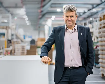 Jens Kriete in dark suit stands beside stacked paper sheets in brightly lit industrial production warehouse.