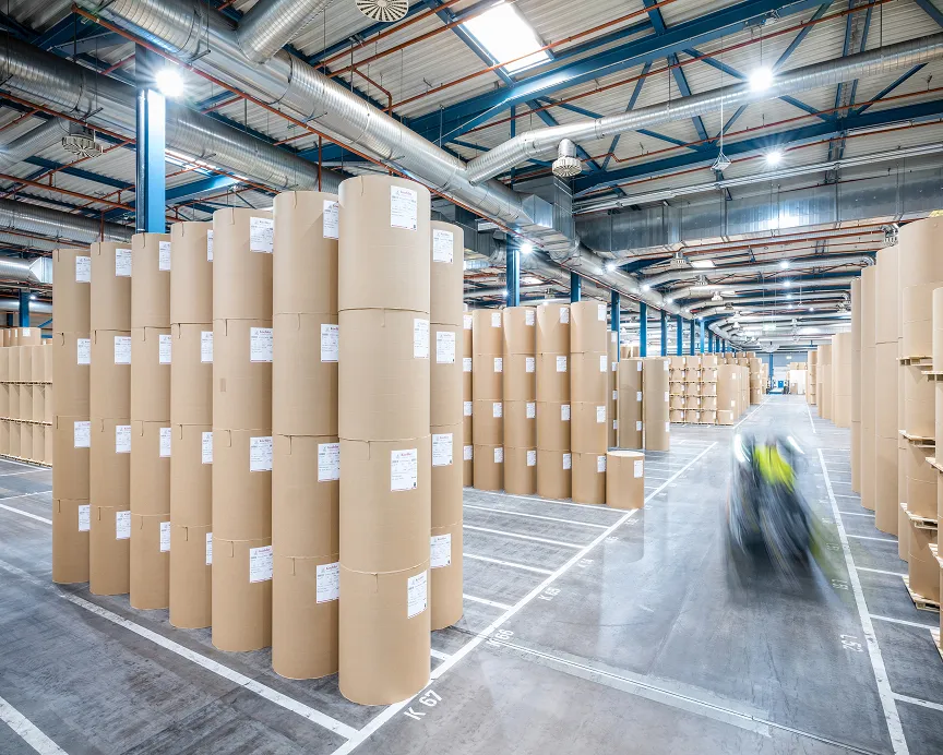 Large warehouse storing tall stacks of wide paper reels, marked driving lanes and blurred forklift moving through aisle.