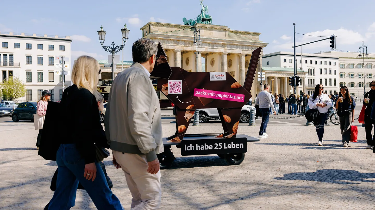 People walking past  origami cat figure with QR code and packs-mit-papier.faz.net banner, Brandenburg Gate in background