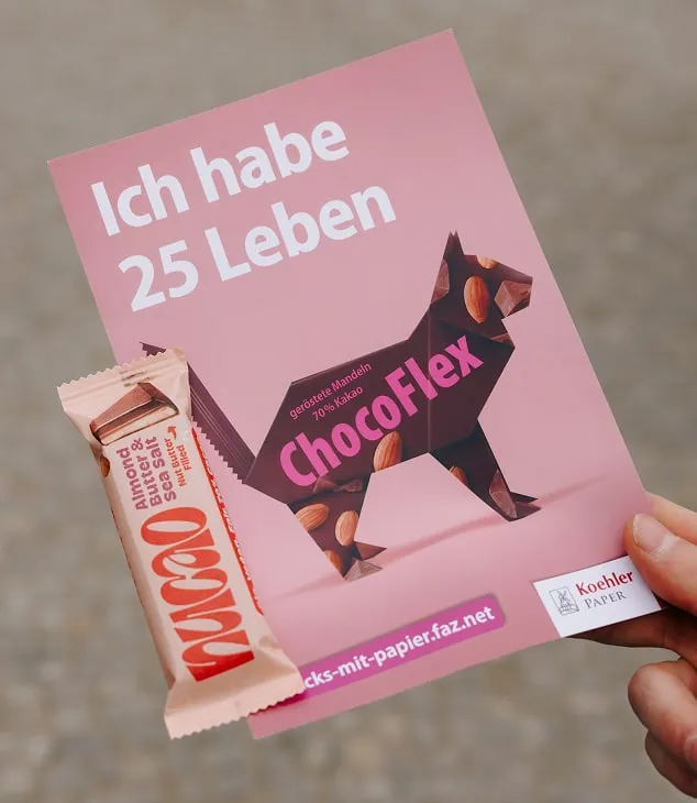Hand holding pink flyer for "Pack's mit Papier" campaign with "Ich habe 25 Leben" slogan, fronted by Nucao Almond Butter & Sea Salt bar
