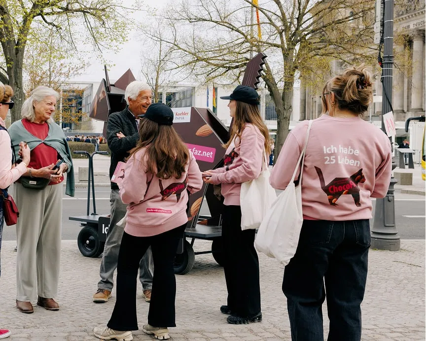 Promo team in pink campaign sweaters with slogan "Ich habe 25 Leben" talking to pedestrians beside origami cat figure on cart