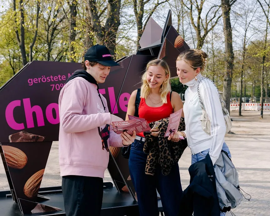 A young promoter in a pink hoodie hands two women flyers for the &ldquo;Pack's mit Papier&rdquo; campaign, featuring the slogan &ldquo;I have 25 lives,&rdquo; in front of a large origami cat